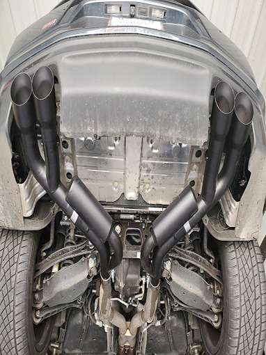 Underside view of a car's dual exhaust system; two black, V-shaped pipes extend from the vehicle's undercarriage, showcasing a custom aftermarket modification.