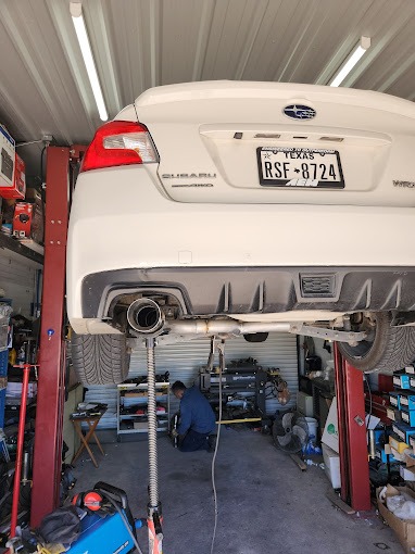 White Subaru WRX raised on a car lift in an auto repair shop. A mechanic works underneath, performing maintenance or repairs.