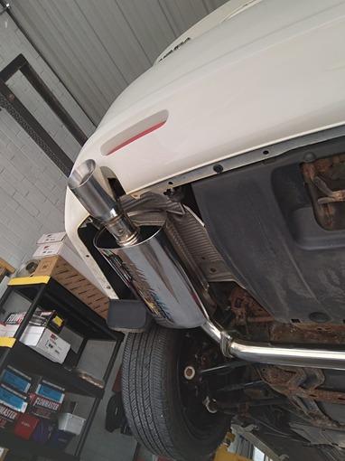 Undercarriage view of a white car in a garage, showcasing a newly installed chrome exhaust pipe. The polished stainless steel muffler is prominently displayed.