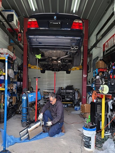 Mechanic installing a new dual exhaust on a raised BMW in a busy auto repair shop. The mechanic is kneeling, working on the underside of the vehicle.